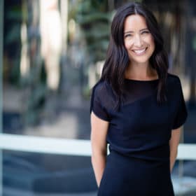 Professional headshot of a woman in a sleek black dress, smiling confidently in a modern, blurred outdoor setting. This image showcases an approachable and polished appearance, ideal for corporate profiles or professional reviews.