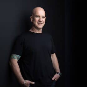 Portrait of a bald man in a casual black t-shirt, leaning confidently against a dark background with subtle lighting highlighting his relaxed yet professional demeanor. His tattooed arm adds a touch of personality and individuality.