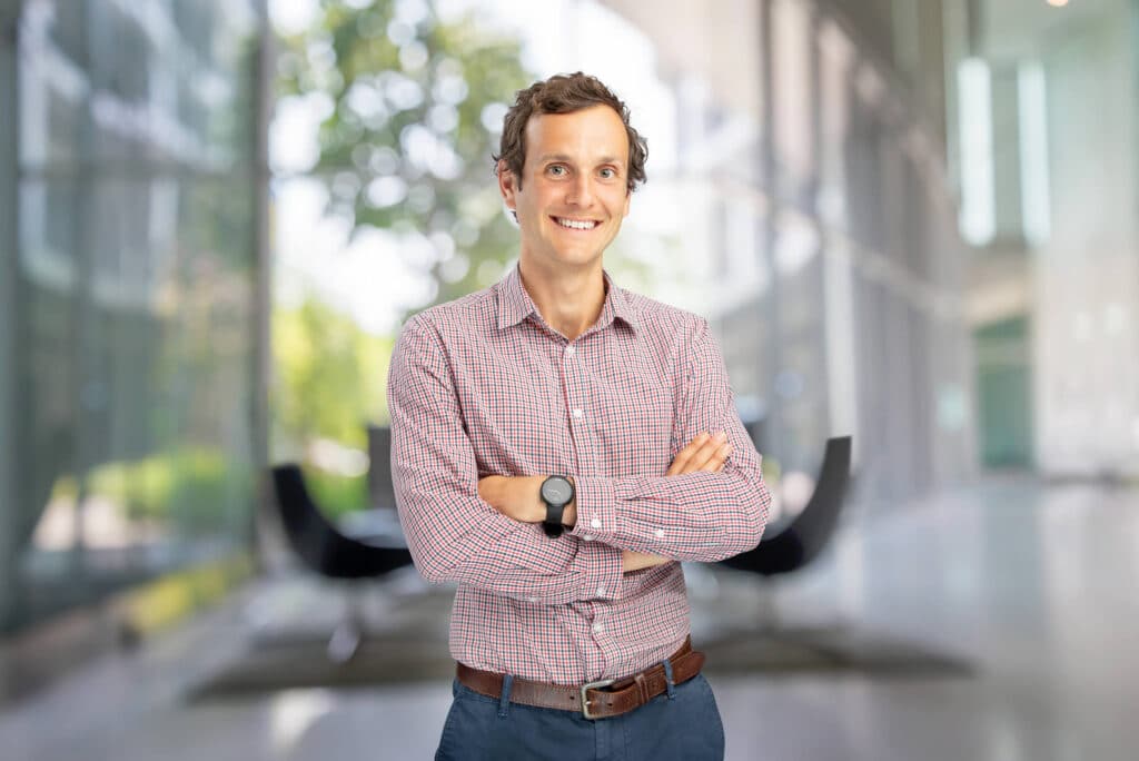 Professional corporate headshot of a smiling man in a checkered shirt, blue pants, and a brown belt, standing with arms crossed in a modern office setting featuring large glass windows and a blurred background of green foliage and sleek furniture. Captured by Hero Shot Photography, showcasing a polished and approachable corporate image.