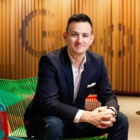 Professional portrait of a confident man in a tailored blazer sitting in a modern office setting with a stylish wooden panel backdrop and vibrant contemporary seating. The image portrays sophistication, success, and a polished business presence ideal for 5-star reviews or professional recommendations.