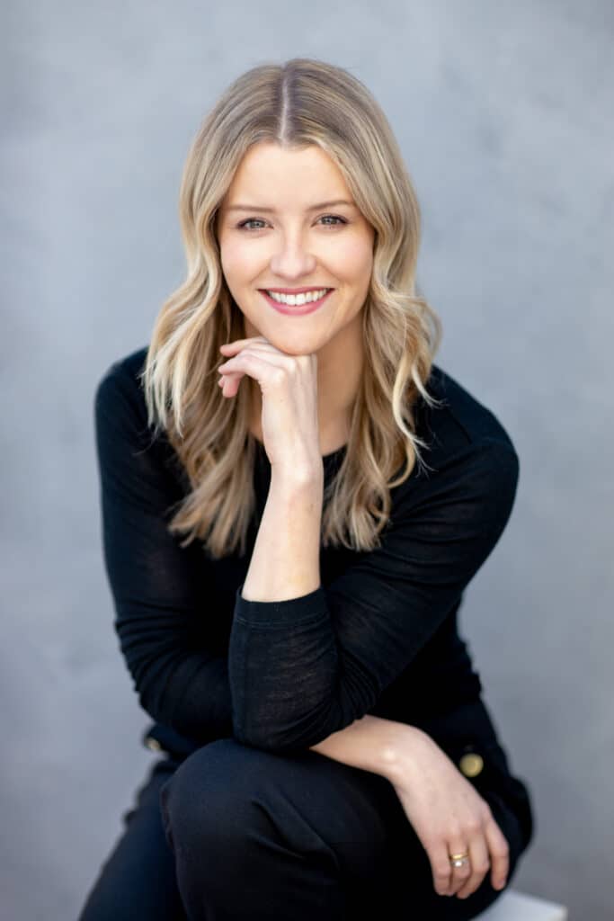 Professional individual headshot of a smiling woman with blonde wavy hair, wearing a black outfit, sitting with her chin resting on her hand. Captured outdoors against a soft, neutral background, exuding warmth, confidence, and approachability. A perfect blend of natural light and professional photography by Hero Shot Photography.