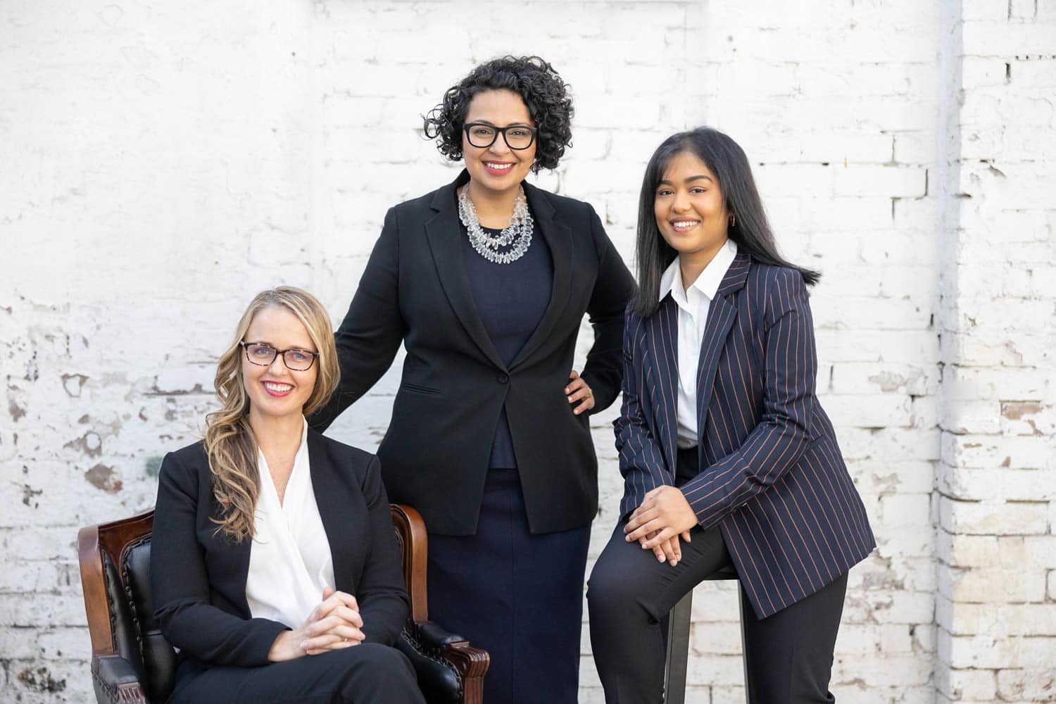 Professional group portrait of three women in formal business attire, posed against a rustic white brick wall. Captured by Hero Shot Photography, showcasing diversity, confidence, and professionalism.