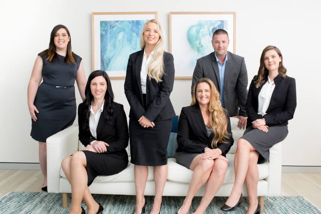 Professional group photo of a business team in formal attire, seated and standing around a white sofa, captured in a modern office setting with framed abstract artwork and clean decor. Captured by Hero Shot Photography, highlighting teamwork, professionalism, and approachability.