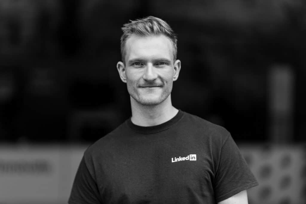 Black and white headshot of a young man wearing a LinkedIn-branded t-shirt, smiling subtly while standing against a blurred dark background. His clean-cut hairstyle and relaxed expression convey professionalism and confidence, ideal for corporate and professional branding.