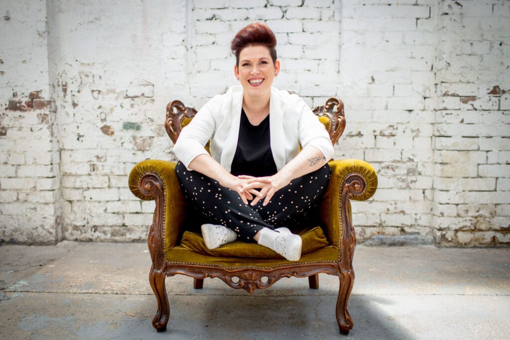 Personal branding photograph of a confident woman sitting cross-legged on a vintage mustard-yellow armchair, set against a rustic white brick wall. She wears a white blazer, black top, polka-dot trousers, and casual sneakers, exuding a blend of professionalism and approachability. The natural lighting enhances her vibrant smile and tattoo detail, reflecting individuality and creativity.