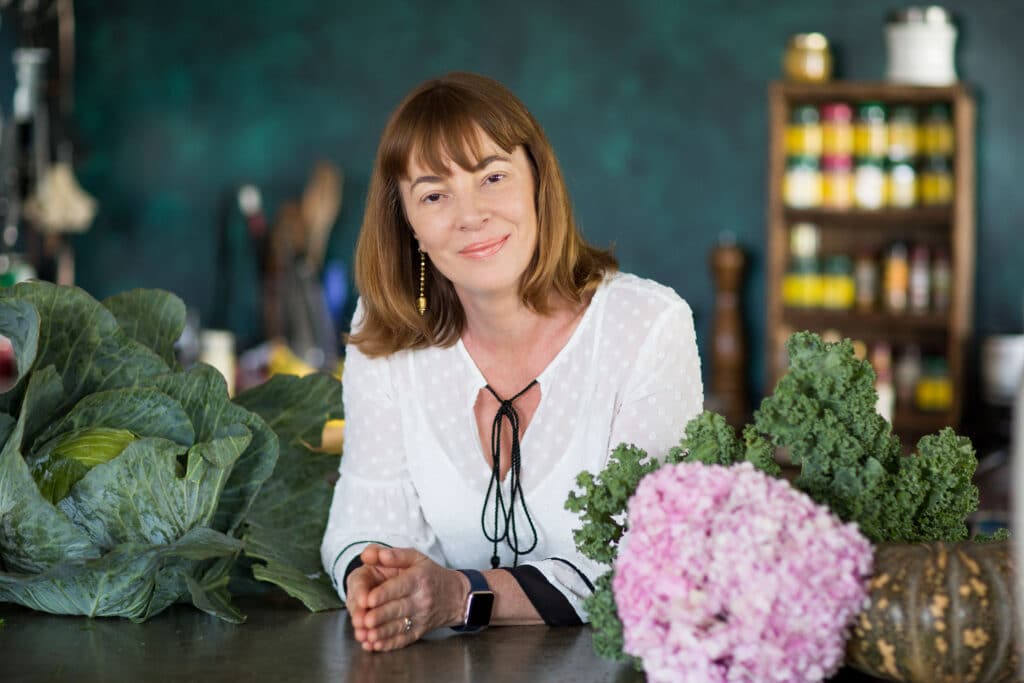 A personal branding photograph of a woman in a cozy kitchen setting, leaning on a counter surrounded by fresh produce, including leafy greens, kale, and a pink hydrangea bouquet. She wears a white blouse with delicate polka dots and gold earrings, exuding a calm and approachable demeanor. The background features a rustic shelf stocked with colorful jars and spices, adding warmth and vibrancy to the scene.