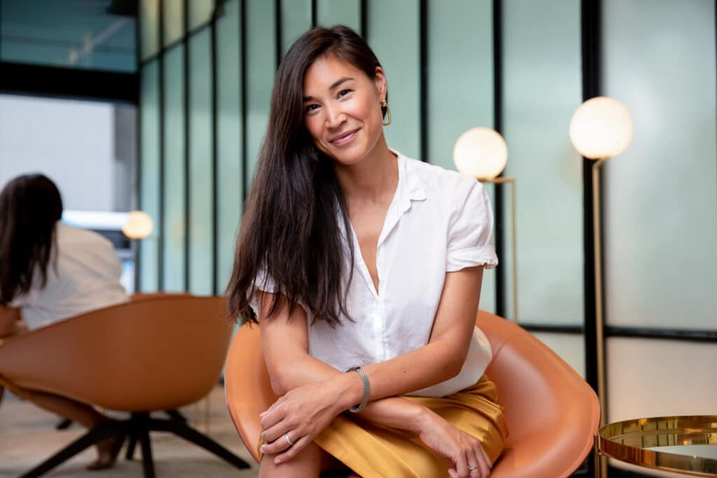 Professional portrait of a smiling woman with long dark hair, seated on a modern orange chair in a stylish office setting with soft lighting and a glass wall background. Captured by Hero Shot Photography, emphasizing confidence, creativity, and professionalism.