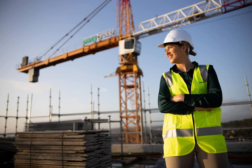 Professional business branding photograph of a female construction professional at a worksite captured by Hero Shot Photography. She is wearing a high-visibility yellow vest and a white hard hat with 'Piety Group' branding, confidently standing with crossed arms against the backdrop of a construction crane and stacked building materials. The golden hour sunlight highlights her empowering stance and the dynamic construction environment.