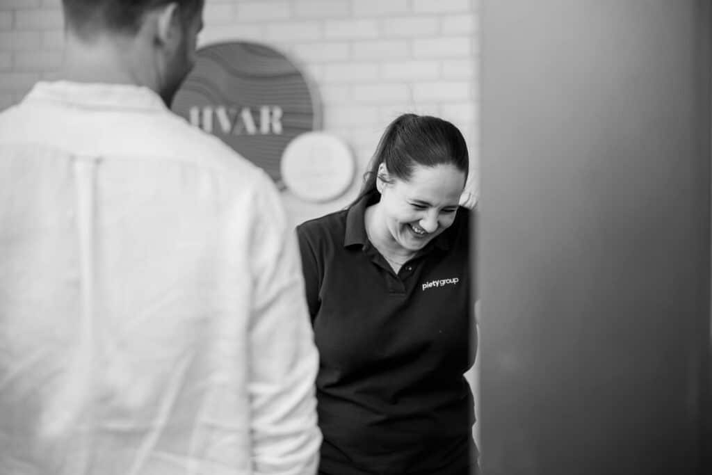 Black-and-white business branding photograph captured by Hero Shot Photography, featuring a joyful moment of a professional woman wearing a branded polo shirt with 'Piety Group' embroidery. She is caught laughing naturally in a casual office environment, interacting with a colleague blurred in the foreground. The composition exudes authenticity, approachability, and team collaboration, ideal for showcasing company culture and values.