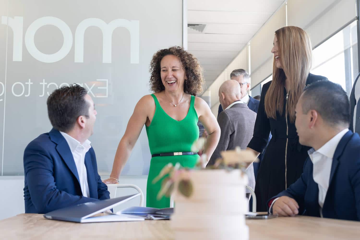 Casual office interaction featuring a smiling woman in a green dress and her colleagues in business attire, engaged in conversation around a table with documents and a decorative plant. Captured in a bright, modern office setting by Hero Shot Photography.