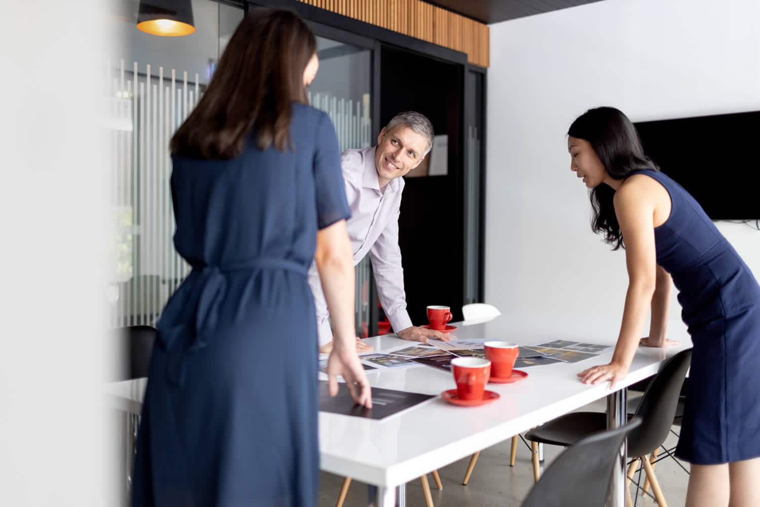 Office team collaboration with two women and one man engaged in discussion around a white table with red coffee cups, in a modern workspace featuring glass walls and wooden accents. Captured by Hero Shot Photography, highlighting teamwork, creativity, and a professional atmosphere.