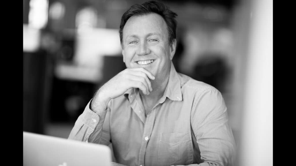 Black and white professional headshot of a smiling man seated at a desk, leaning on one hand with a relaxed and approachable demeanor. He is wearing a casual button-down shirt, and a blurred office background provides a modern yet understated setting.