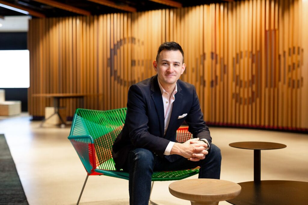 Professional portrait of a confident man seated on a modern green and red chair in a contemporary office space with a Google-branded wooden backdrop. Captured by Hero Shot Photography, showcasing professionalism, creativity, and approachability.