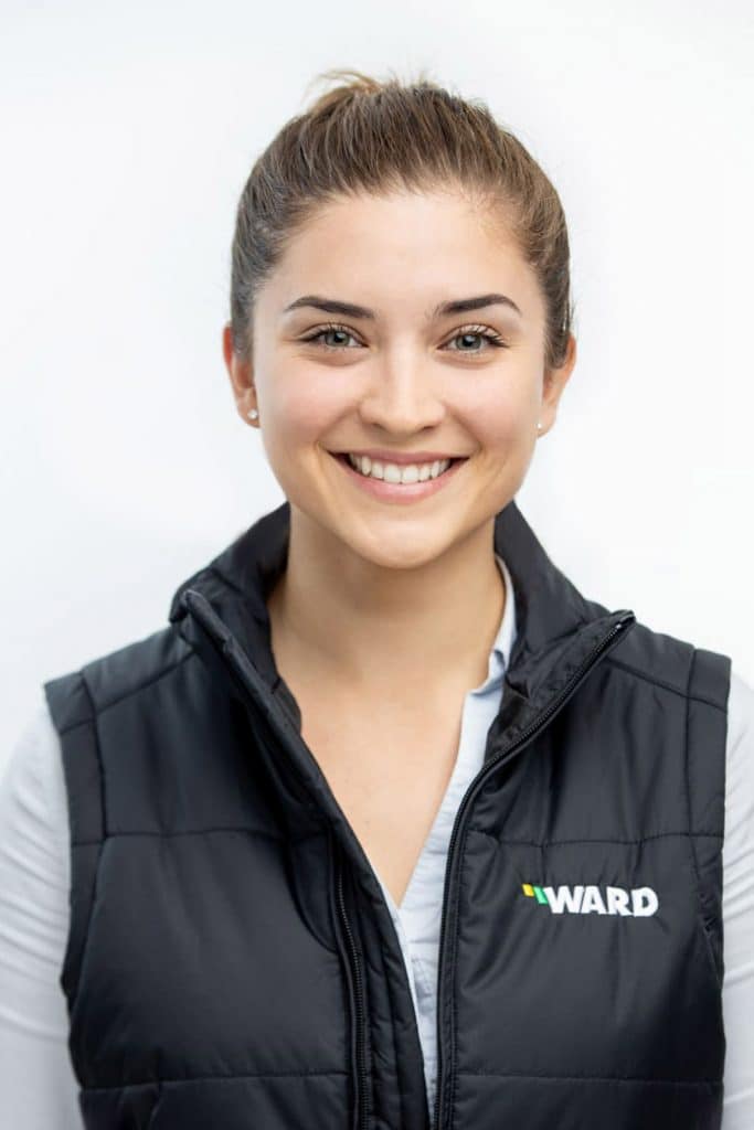 Professional headshot of a young woman with her hair tied back in a neat bun, wearing a black quilted vest with the 'Ward' logo over a light blue collared shirt. She exudes confidence and approachability, captured against a clean, bright background that highlights her friendly smile and sharp professional appearance.