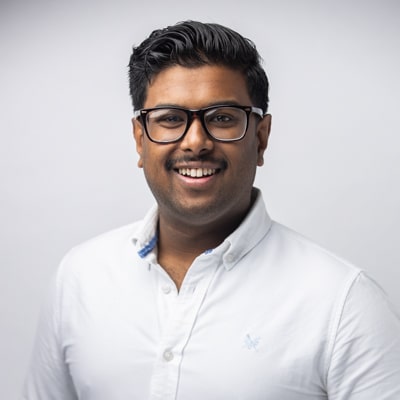 Professional portrait of a smiling man with short dark hair, wearing glasses and a crisp white collared shirt, captured against a soft gray background by Hero Shot Photography. Highlights confidence, professionalism, and approachability.