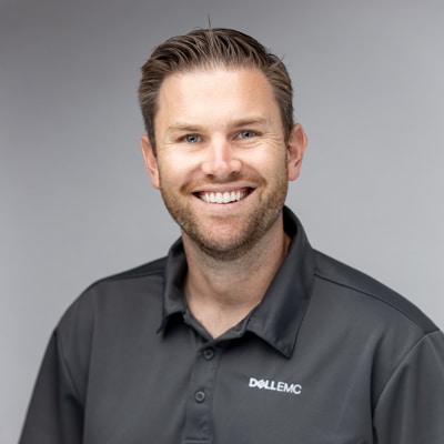 Professional portrait of a smiling man wearing a gray Dell EMC polo shirt, captured against a soft gray background by Hero Shot Photography. Highlights confidence, professionalism, and approachability, ideal for corporate branding and team profiles.