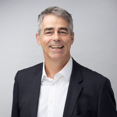 Professional portrait of a smiling man with salt-and-pepper hair, wearing a black blazer and white shirt, captured against a soft gray background by Hero Shot Photography. Highlights professionalism, confidence, and approachability.