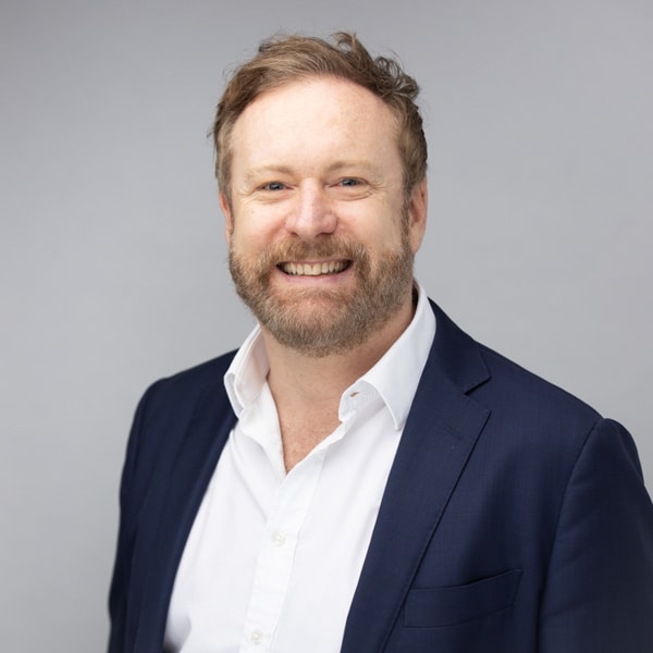 Professional portrait of a smiling man with light hair and a beard, dressed in a navy suit and white shirt, captured against a soft gray background by Hero Shot Photography. Highlights confidence, warmth, and professionalism for corporate and branding purposes.