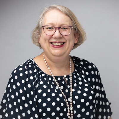 Professional portrait of a smiling woman with short blonde hair, wearing glasses, a polka-dot blouse, and a pearl necklace, captured against a soft gray background by Hero Shot Photography. Highlights approachability, warmth, and professionalism.