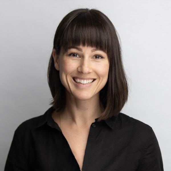 Professional portrait of a smiling woman with shoulder-length dark hair and bangs, wearing a black collared shirt, captured against a light gray background by Hero Shot Photography. Highlights confidence, warmth, and professionalism for personal branding and corporate use.