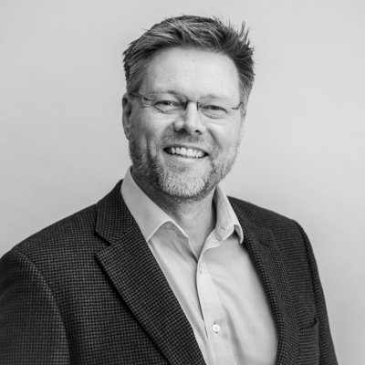 Black-and-white professional portrait of a smiling man wearing glasses, a checkered blazer, and a collared shirt, captured against a light gray background by Hero Shot Photography. Highlights professionalism, confidence, and approachability.