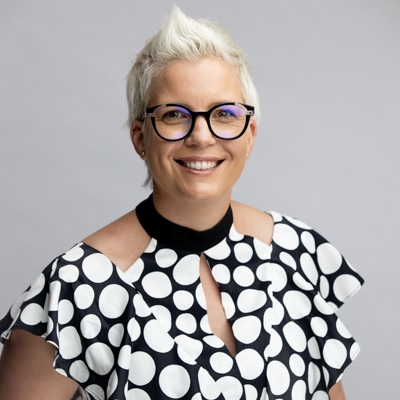 Professional portrait of a smiling woman with short white hair, wearing stylish glasses and a polka-dot patterned top, captured against a soft gray background by Hero Shot Photography. Highlights confidence, creativity, and individuality.