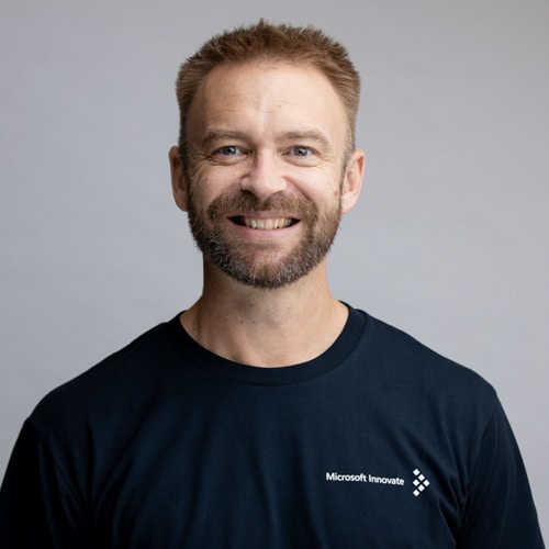 Professional portrait of a smiling man wearing a navy Microsoft Innovate t-shirt, captured against a soft gray background by Hero Shot Photography. Highlights confidence, approachability, and professionalism for corporate branding and team profiles.