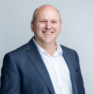 Professional portrait of a smiling man in a navy blue suit and white shirt, captured against a soft gray background by Hero Shot Photography. Exemplifying exceptional headshot photography that highlights confidence, professionalism, and approachability. Ideal for corporate profiles, LinkedIn, and personal branding.