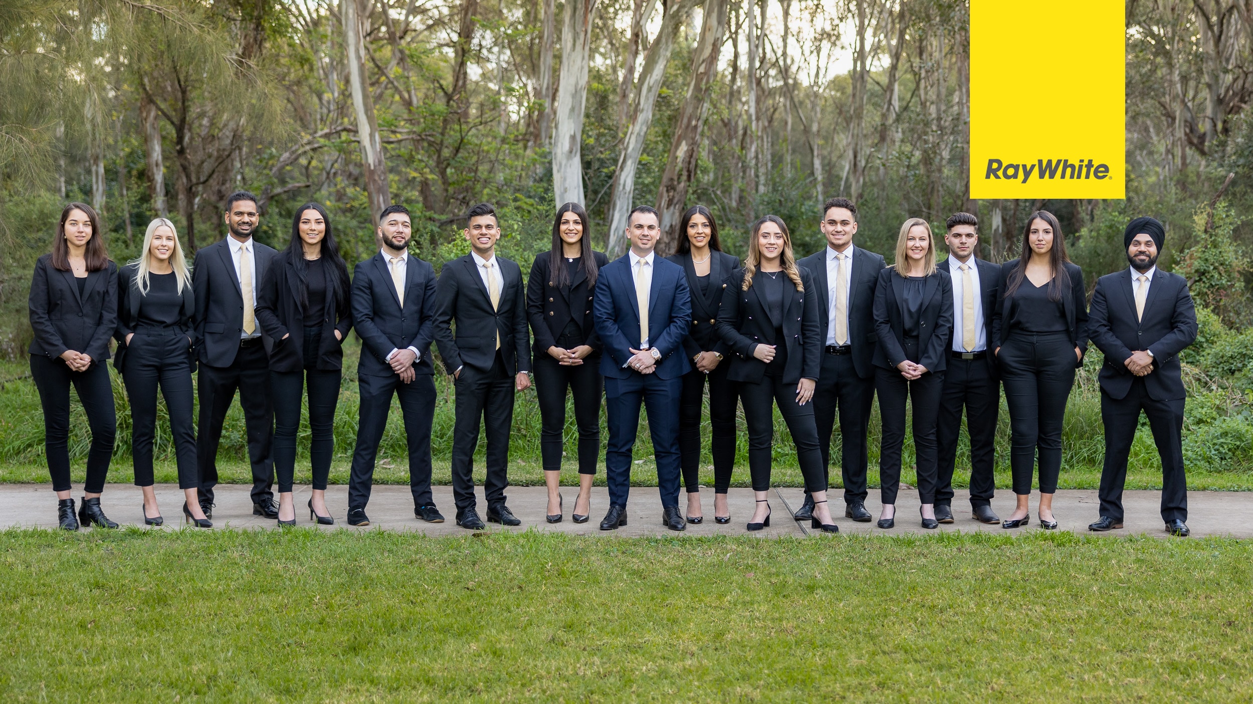 Group photo of the Ray White team standing outdoors in formal business attire with matching black suits and gold ties, set against a serene natural background of trees and greenery. Captured by Hero Shot Photography, showcasing professionalism, unity, and corporate identity.