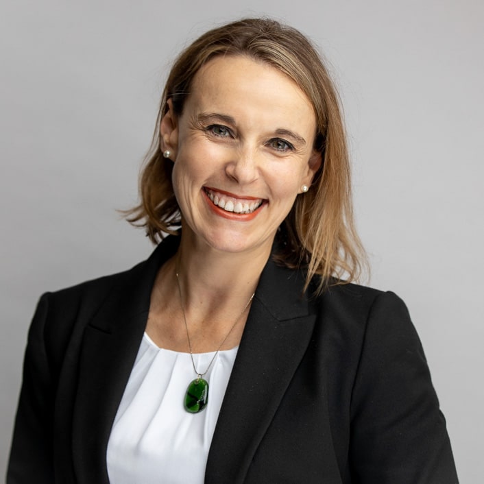 Professional portrait of a smiling woman with shoulder-length blonde hair, wearing a black blazer, white top, and green pendant necklace, captured against a soft gray background by Hero Shot Photography. Highlights confidence, approachability, and professionalism.