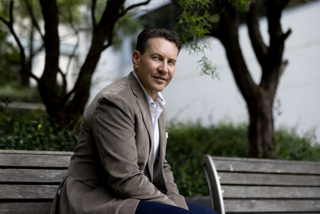 headshot of a man sitting on a park bench with greenery behind